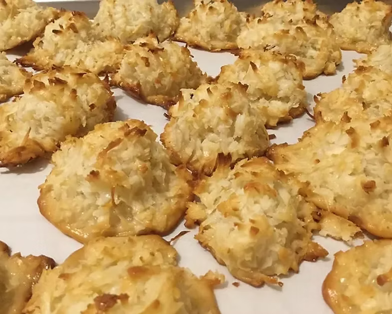 A close-up shot of freshly baked coconut macarons on a white baking sheet, showing their golden-brown toasted coconut tops.